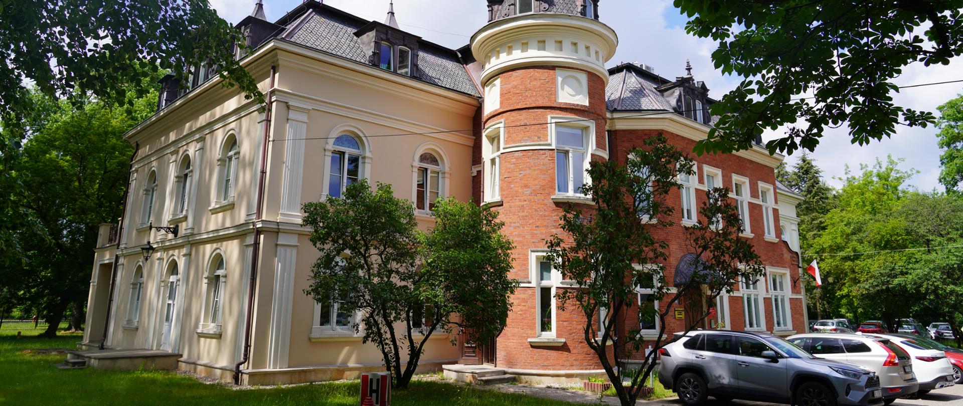 Pałac Hasbacha w Białymstoku. Budynek o jasnej elewacji, z elementami czerwonej cegły, znajdujący się w otoczeniu drzew.