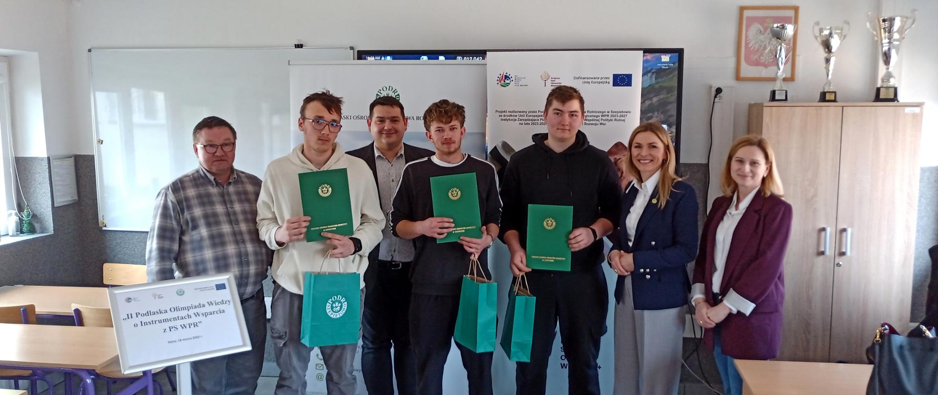 Grupa osób pozuje do zdjęcia, wśród nich trzej uczniowie z dyplomami i nagrodami zdobytymi w konkursie.