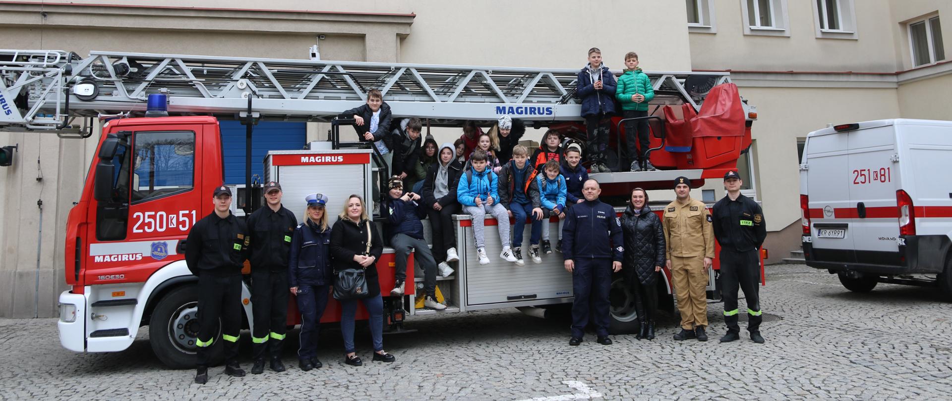  Pamiątkowe zdjęcie uczniów, wychowawców i funkcjonariuszy policji i PSP. Dzieci siedzą i stoją na pojeździe, którym jest drabina mechaniczna. 