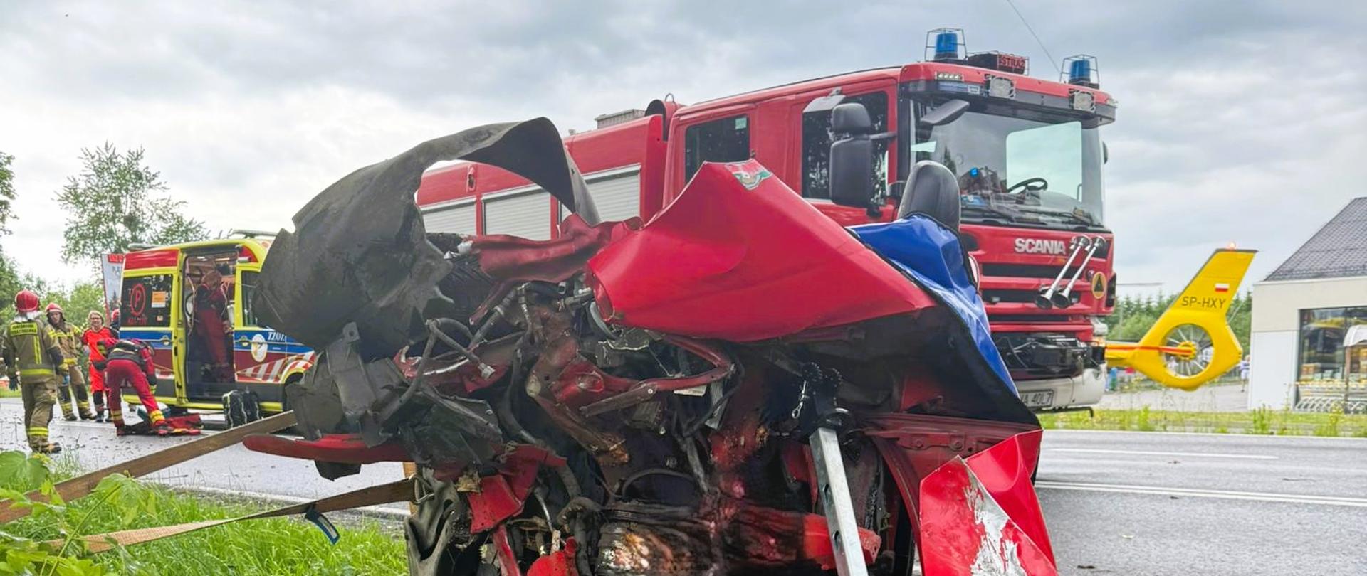 Na środku czerwony mocno zniszczony samochód osobowy ustabilizowany podporami ratowniczymi. W tle samochód pożarniczy. Po lewej stronie w tle karetka.