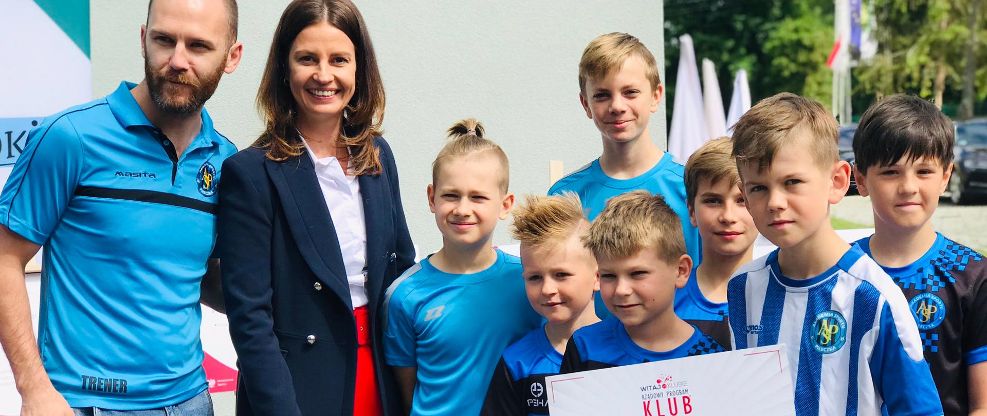 Danuta Dmowska-Andrzejuk z dziećmi w sportowych strojach trzymających bon "Witaj w Klubie"