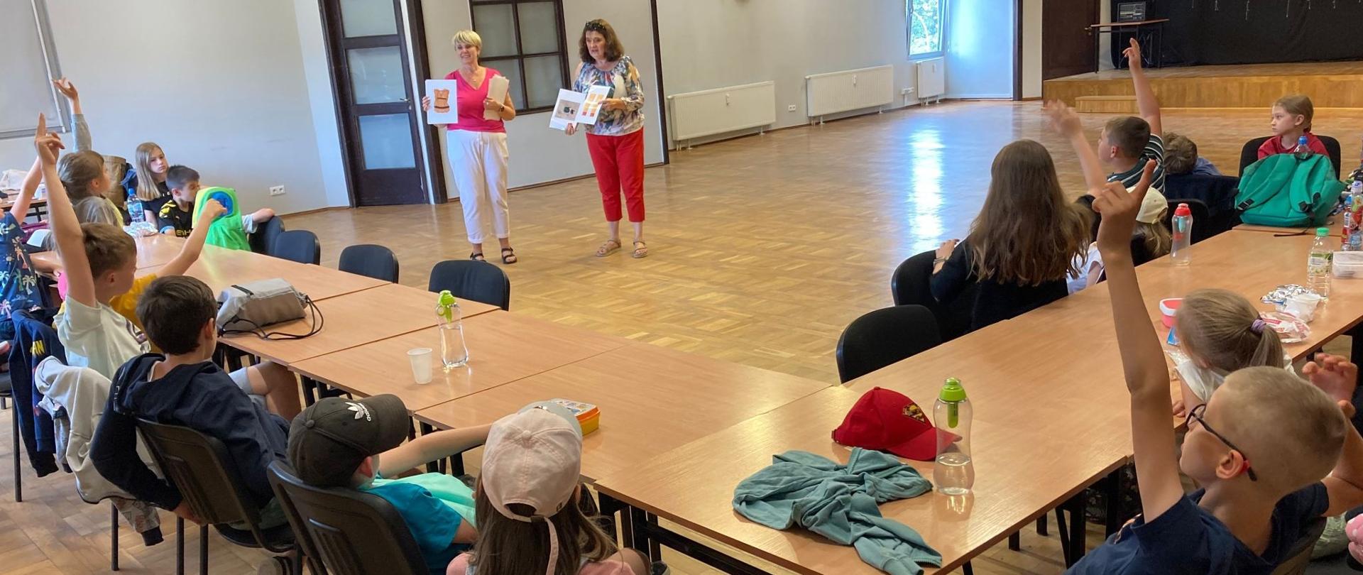 W dużej sali, przy długim stole siedzą dzieci i słuchają dwóch prelegentek, które stoją na środku prezentując plansze edukacyjne 