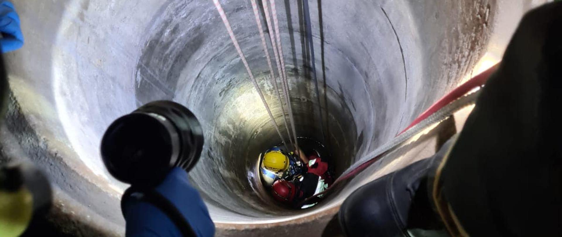 Na zdjęciu znajduje się studnia, wewnątrz jest strażak opuszczony na linach. Wnętrze studni jest oświetlane