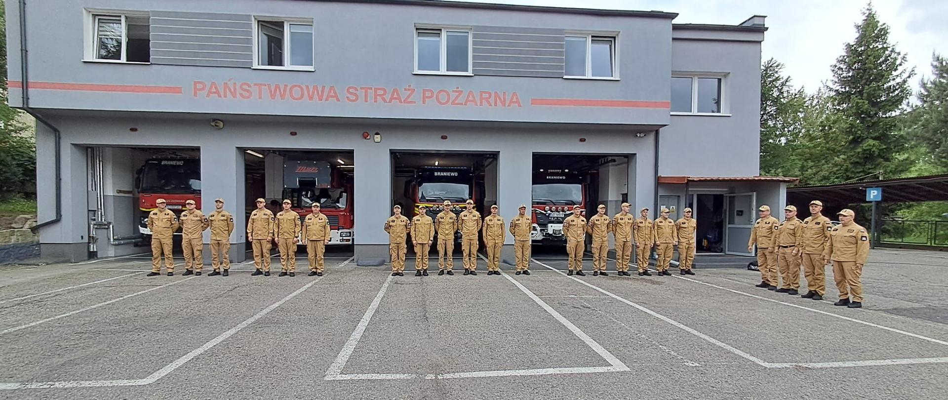 Przed remizą jednostki ratowniczo gaśniczej stoją obok siebie strażacy w jasnych mundurach., 22 strażaków. Otwarte bramy garażowe a w nich samochody strażackie.