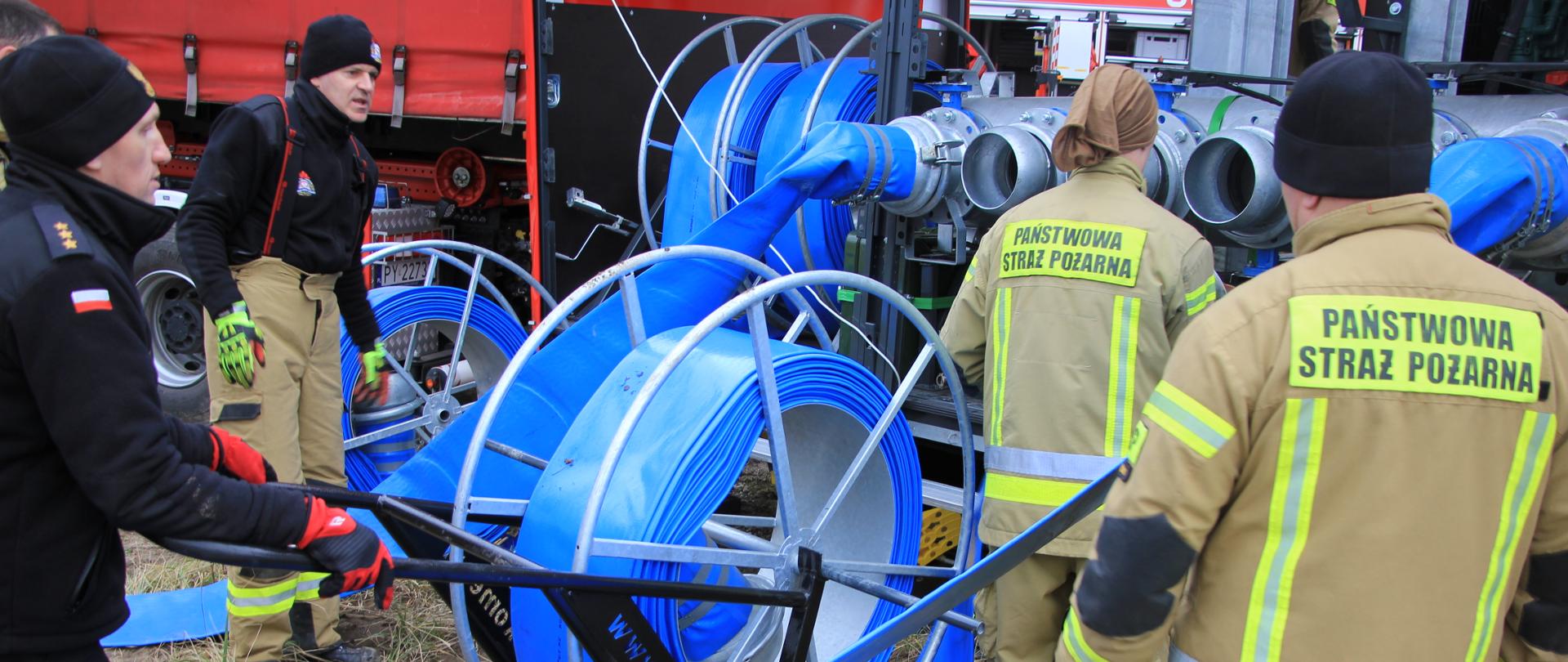 Strażący podłączają węże do mompy na przyczepie.