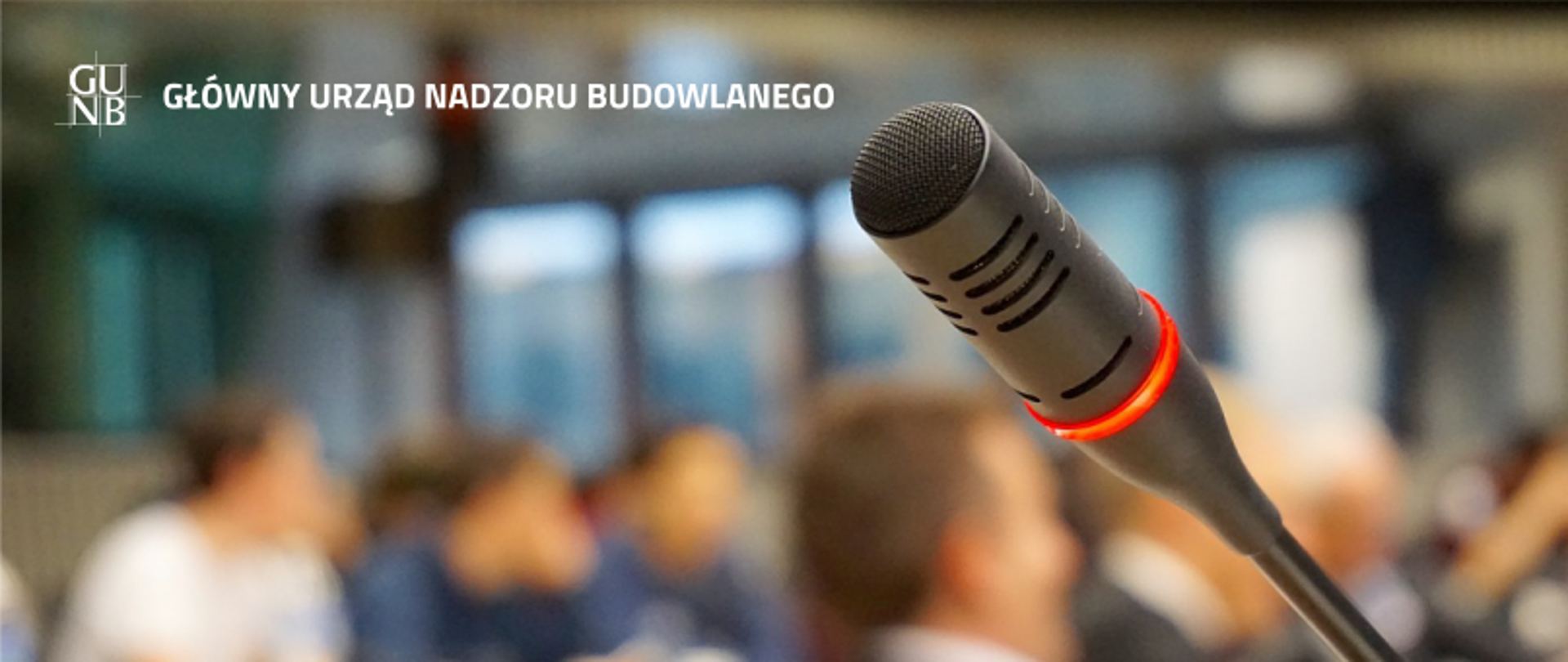 Zbliżenie na włączony mikrofon z czerwoną obwódką, w tle rozmyte sylwetki uczestników spotkania, logo GUNB i napis „GŁÓWNY URZĄD NADZORU BUDOWLANEGO”.
