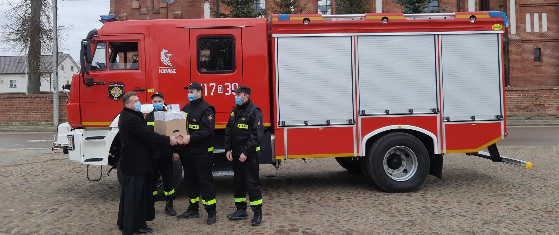 Strażacy przekazują maseczki ochronne księdzu. Zdjęcie wykonane przy wozie strażackim.