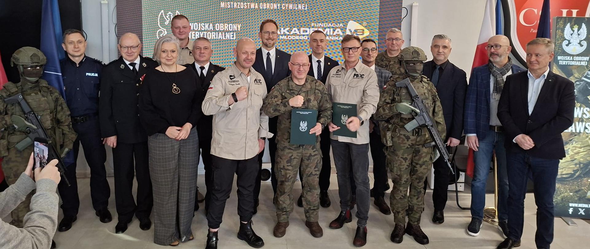 Zaproszeni goście stoją w grupie, z tyłu widoczny banner z napisem moc, który zawieszony jest na szarej ścianie