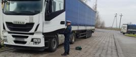 Inspektor ITD stoi obok otwartych drzwi ciężarówki od strony kierowcy i kontroluje dokumenty. Na ziemi, obok rury wydechowej ciągnika siodłowego, stoi dymomierz. 