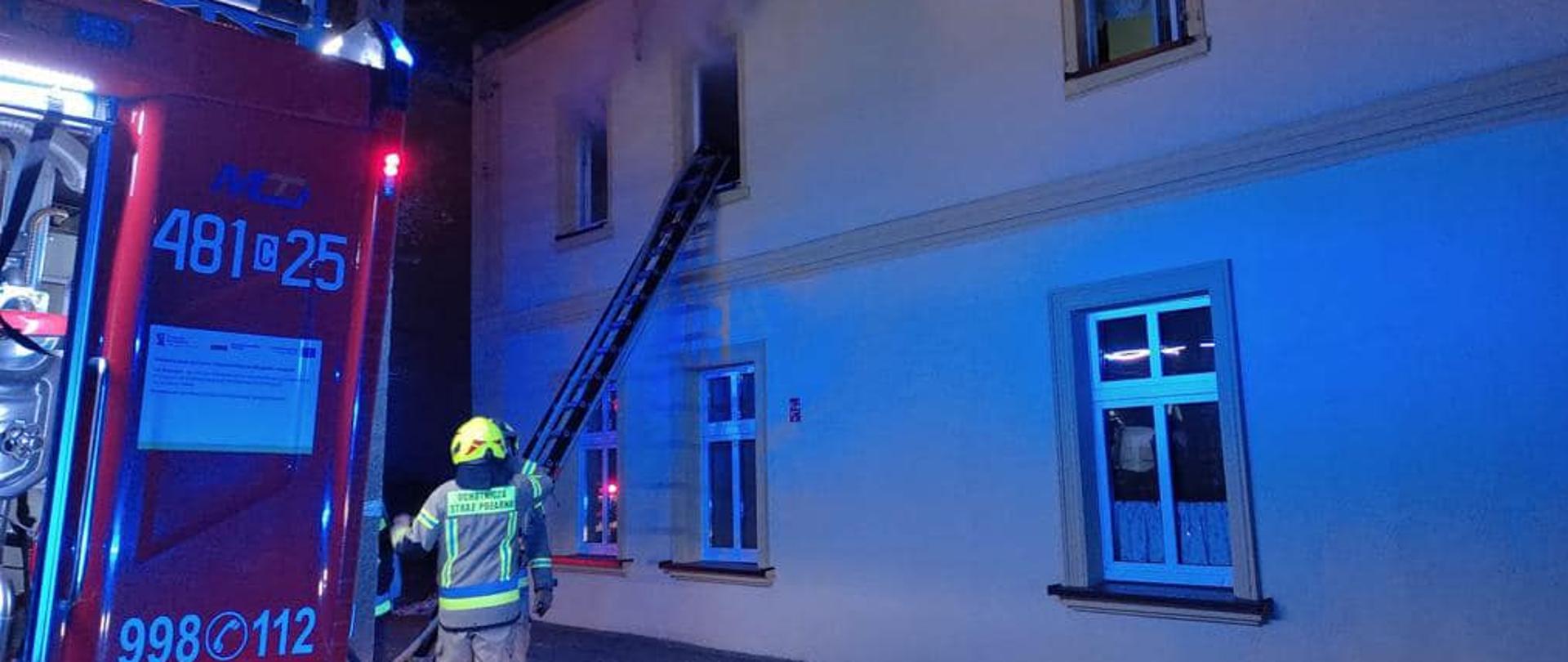 Na pierwszym planie budynek mieszkalny, z którego okien wydostaje się dym. Po lewej stronie wóz strażacki a obok niego strażacy. 