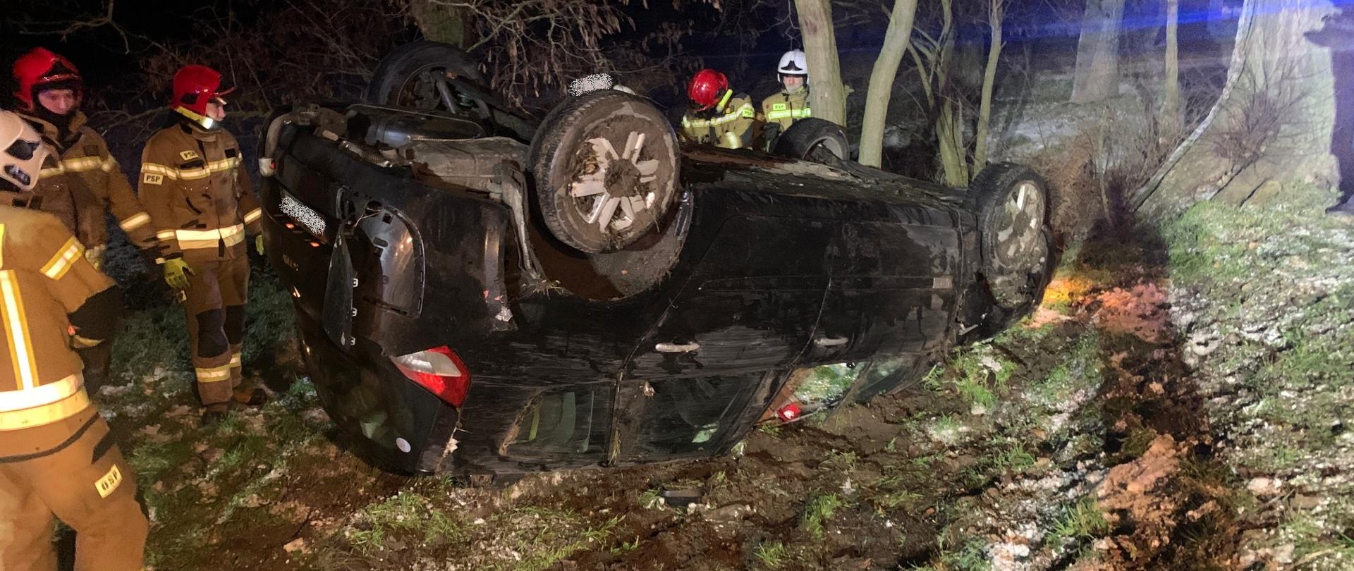 Samochód osobowy koloru czarnego leży na dachu w przydrożnym rowie. Obok stoją strażacy