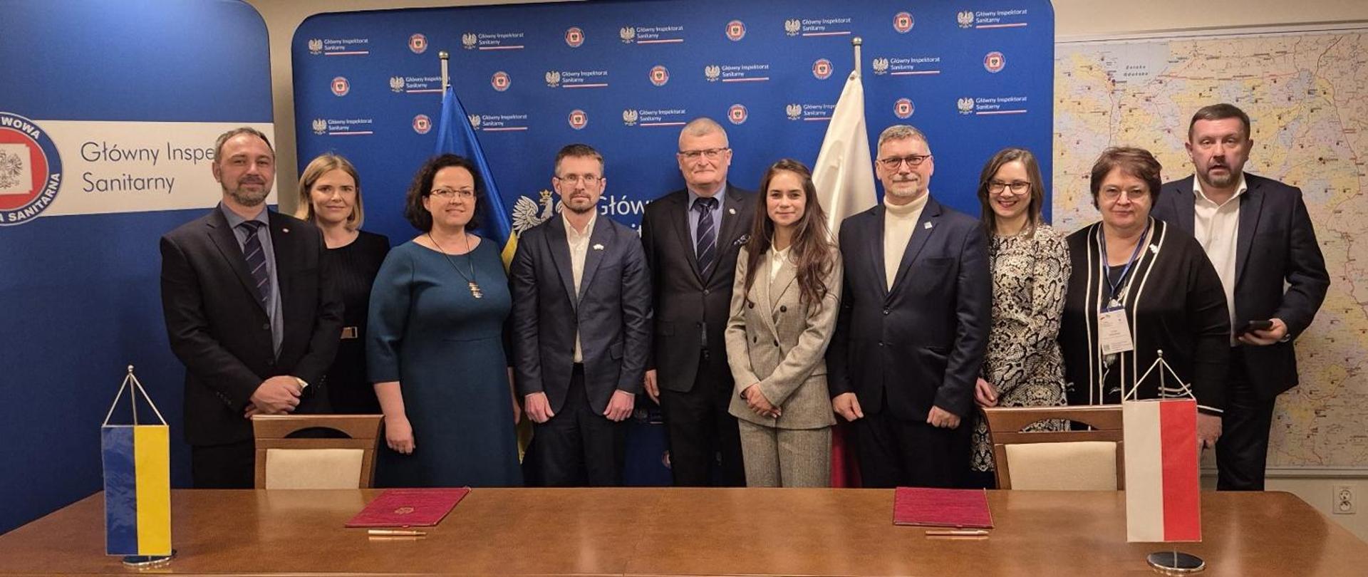 Na zdjęciu widać grupę kilkunastu osób stojących w dwóch rzędach podczas oficjalnego wydarzenia. Tłem jest ściana z niebieskimi panelami zawierającymi oznaczenia Główny Inspektorat Sanitarny.
Przed uczestnikami ustawiono stół, na którym znajdują się dwa dokumenty oraz małe stojaki z flagami Polska i Ukraina.
Osoby na zdjęciu są elegancko ubrane — głównie w garnitury, żakiety i formalne stroje. Wszyscy pozują do zdjęcia, stojąc blisko siebie i uśmiechając się, co sugeruje podpisanie porozumienia, spotkanie robocze lub wspólną deklarację współpracy pomiędzy instytucjami obu krajów.
W tle po prawej stronie widać także dużą mapę, co dodatkowo podkreśla urzędowy i organizacyjny charakter wydarzenia.