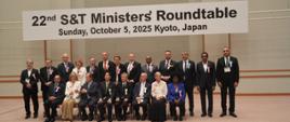 Zdjęcie zbiorowe, pod tablicą z napisem 22nd S&T Ministers'Roundtable stoi grupa kilkunastu osób, przed nimi na ustawionych w rząd krzesłach siedzi osiem osób.