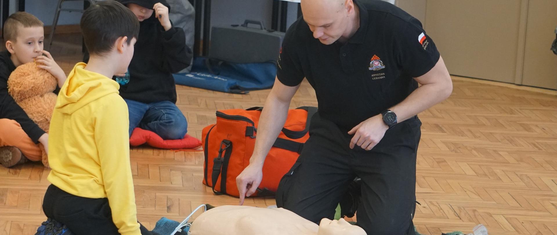 Na zdjęciu znajduje się strażak oraz dzieci. Strażak pokazuje chłopcu jak prowadzić RKO na fantomie.