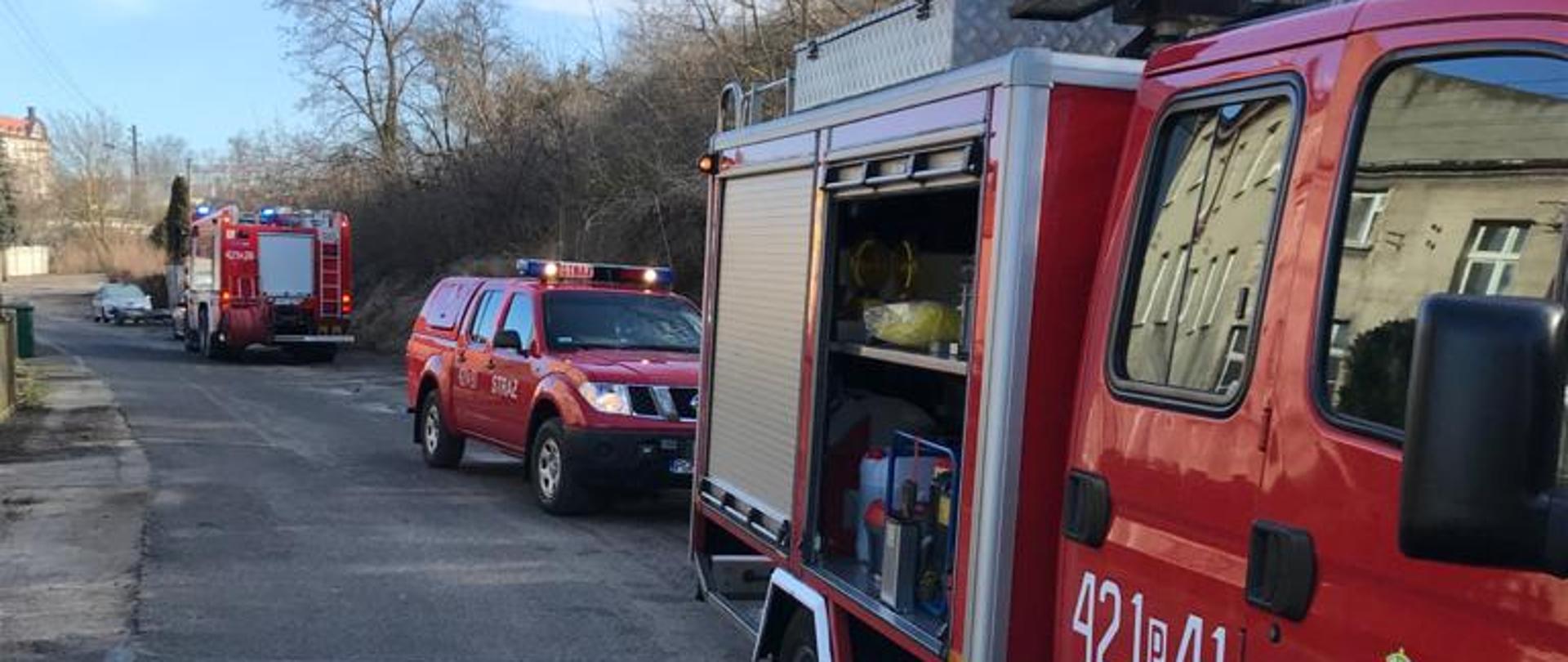 Zdjęcie zrobione w ciągu dnia, pogodnie, niebieskie niebo z białymi chmurami. Na zdjęciu widać trzy samochody gaśnicze zaparkowane jeden za drugim na ulicy. Pierwszy samochód ma otwartą boczną skrytkę na sprzęt