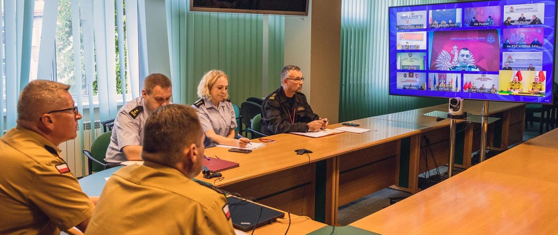 Po lewej stronie pięciu strażaków w umundurowaniu służbowym, siedzących przy biurku i wpatrujących się w monitor stojący po prawej stronie.