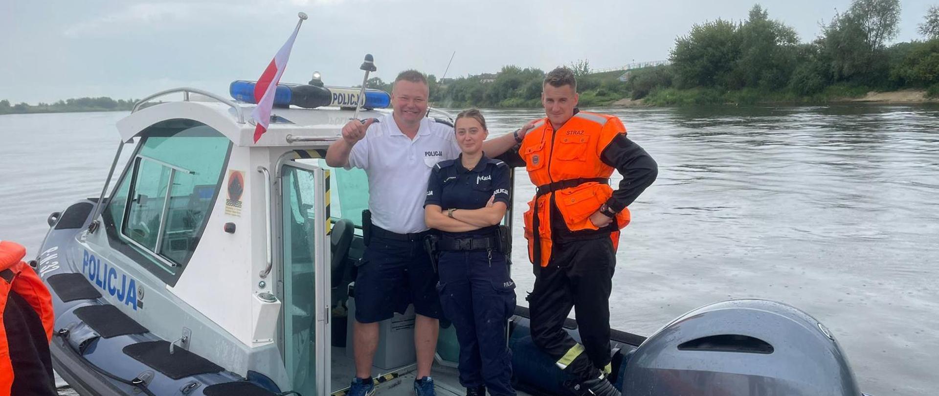 Na motorówce policyjnej stoją trzy osoby - Policjant, Policjantka oraz strażak. Uśmiechają się do zdjęcia.