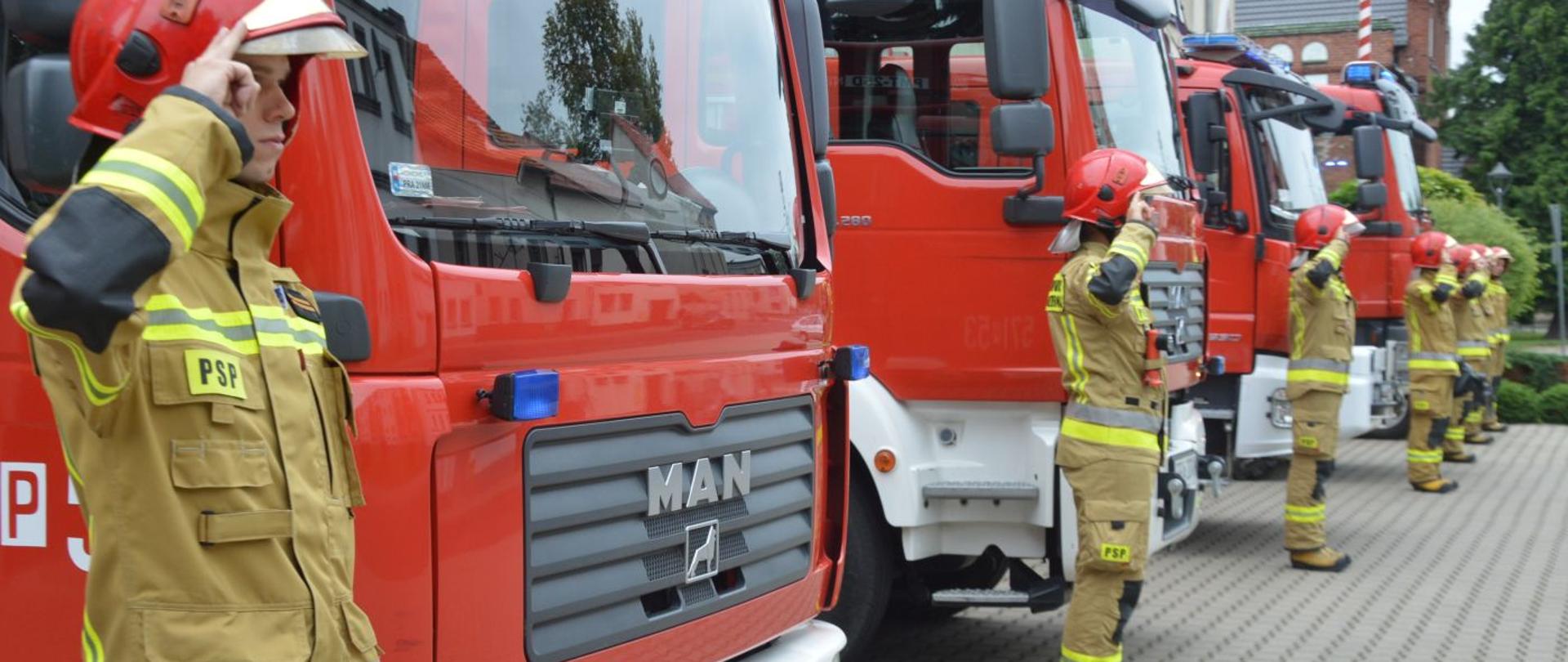Oddanie hołdu powstańcom warszawskim. Plac przed Komendą Powiatową PSP w Rawiczu. Przed bramami garażowymi stoją cztery samochody pożarnicze z włączonymi sygnałami świetlnymi. Przed samochodami w postawie na baczność stoi sześciu strażaków w ubraniach specjalnych i hełmach, którzy salutują.