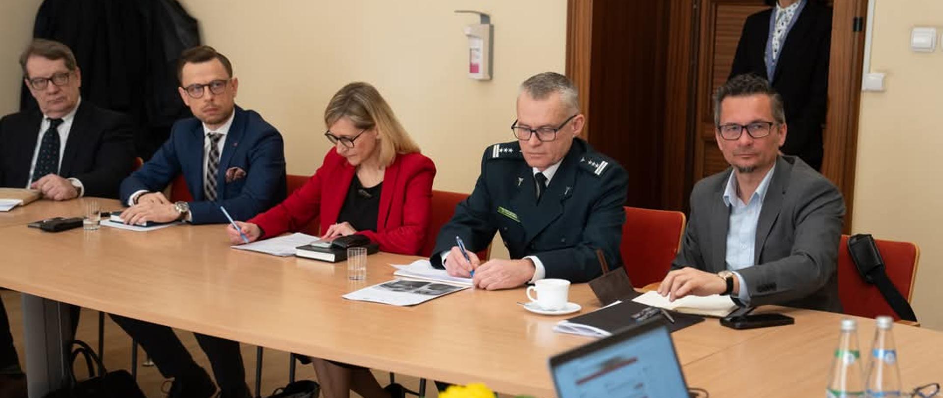 Pięć osób siedzących za długim stołem na spotkaniu w Urzędzie. Na środku siedzi kobieta w czerwonej marynarce, po jej obu stronach po dwóch mężczyzn w garniturach lub mundurach.