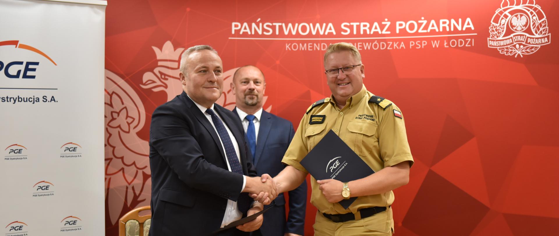 nadbryg. Grzegorz Janowski i Andrzej Piętka - dyrektor generalny PGE Dystrybucja Oddział Łódź, ściskają sobie dłonie, za nimi mężczyzna w garniturze a dalej banery PGE i KW PSP w Łodzi