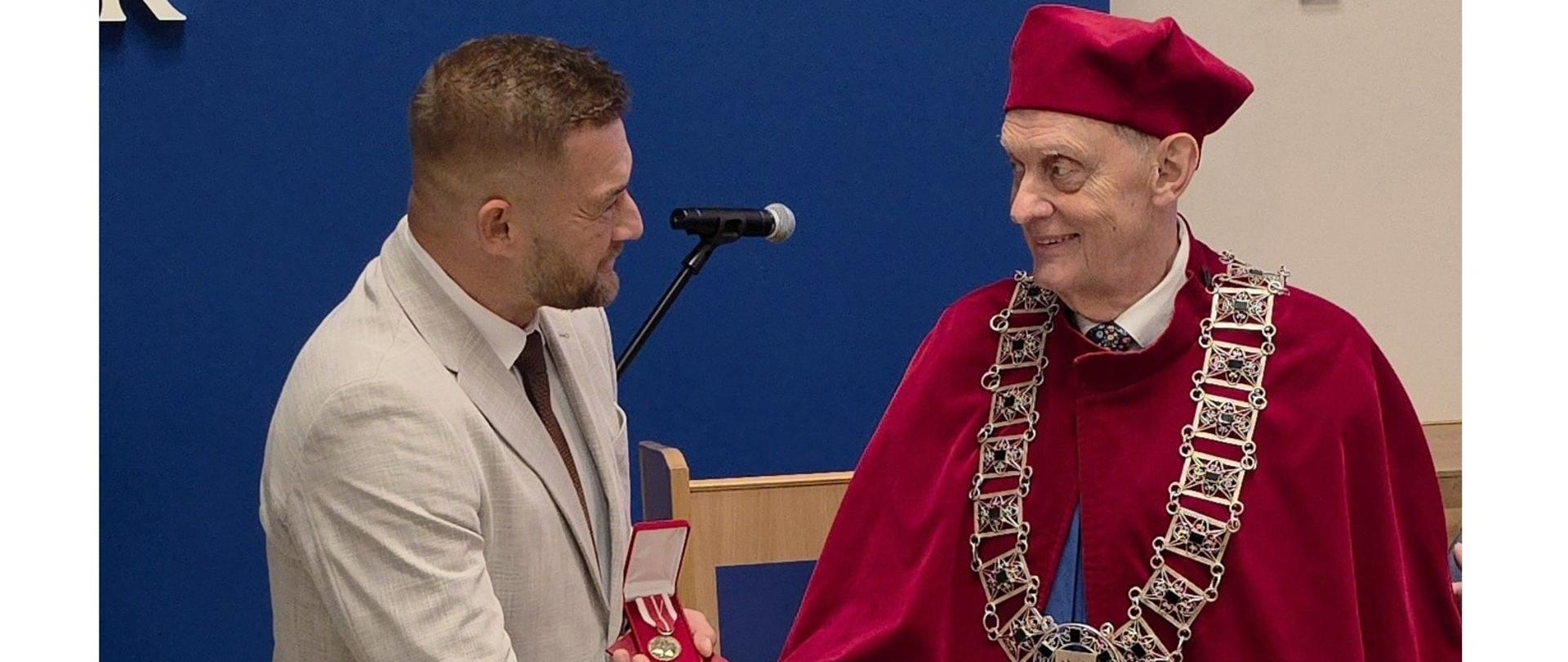 Zdjęcie przedstawia moment wręczenia medalu podczas uroczystości akademickiej. Osoba w jasnym garniturze trzyma otwarte etui z odznaczeniem i ściska dłoń osoby ubranej w bordowy strój ceremonialny z ozdobnym łańcuchem. W tle znajdują się mównica, mikrofon oraz niebieska ściana sali.