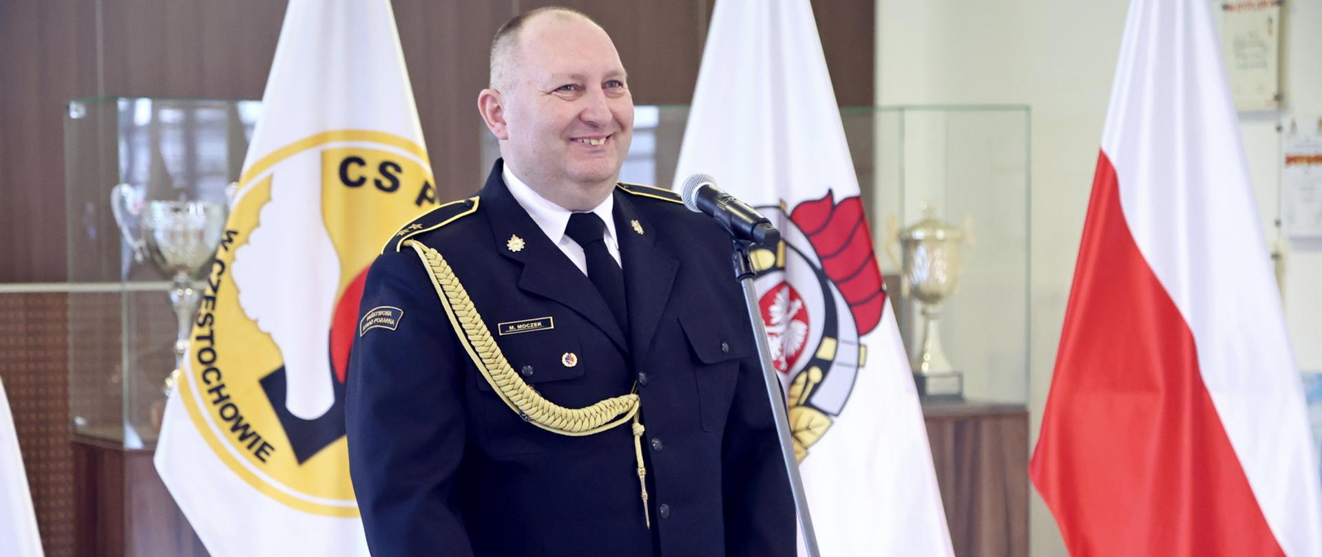 Odchodzący na emeryturę st. asp. przemawiający do mikrofonu, w tle flagi stojące na stojakach