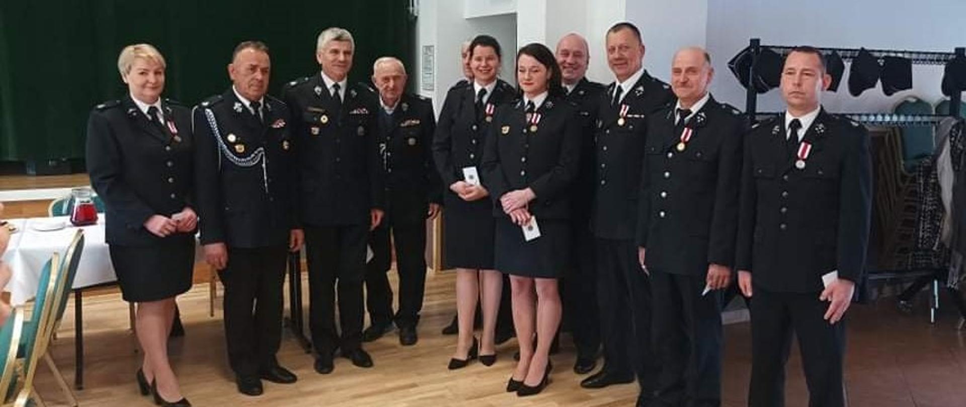 Zdjęcie przedstawia delegatów zarządu gminnego oraz zastępcę komendanta powiatowego w umundurowaniu galowym. Zdjęcie wykonane wewnątrz budynku, z tyłu widać scenę. 