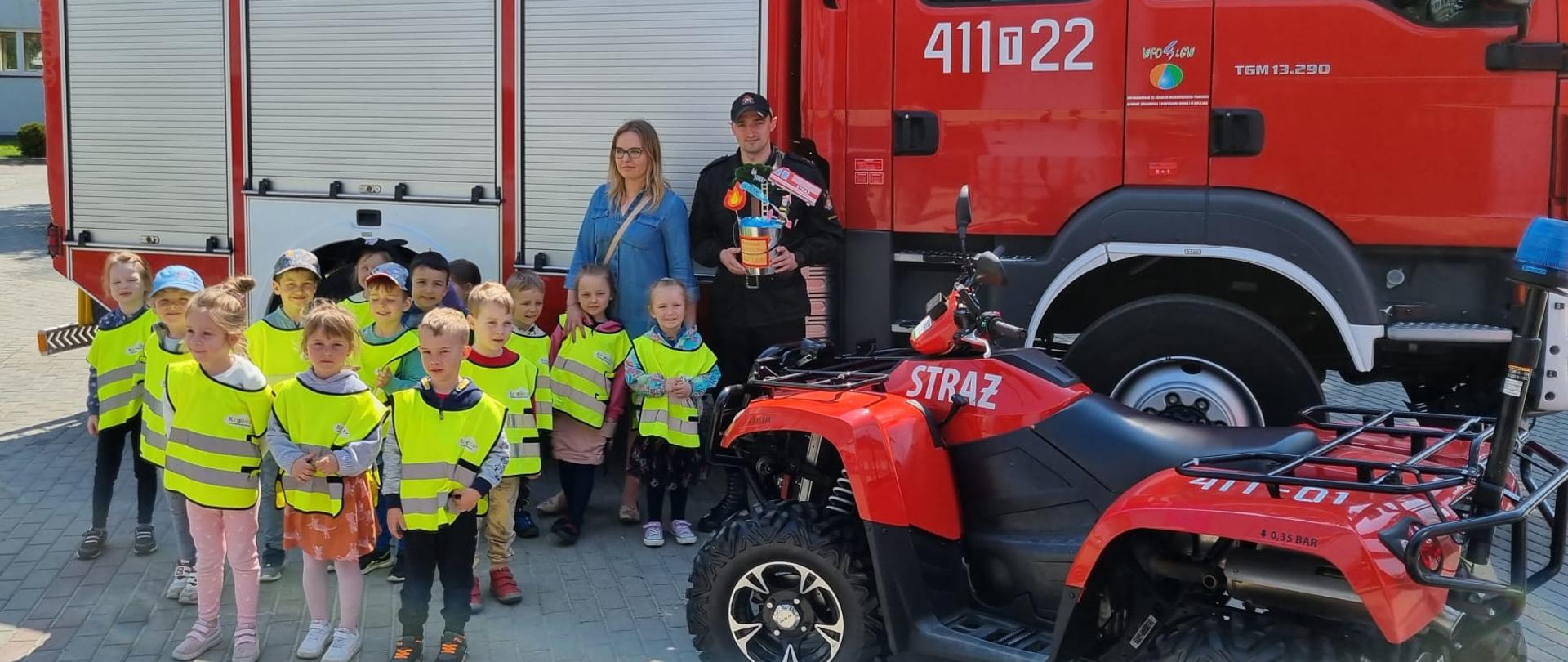 Zdjęcie przedstawia strażaka w umundurowaniu dowódczo-sztabowym, który w rękach trzyma laurkę w kształcie wiaderka z wodą. Obok strażaka znajdują się dzieci w kamizelkach odblaskowych oraz ich opiekunka. Zdjęcie zrobione na zewnątrz na tle samochodu ratowniczo-gaśniczego oraz pojazdu typu quad. 