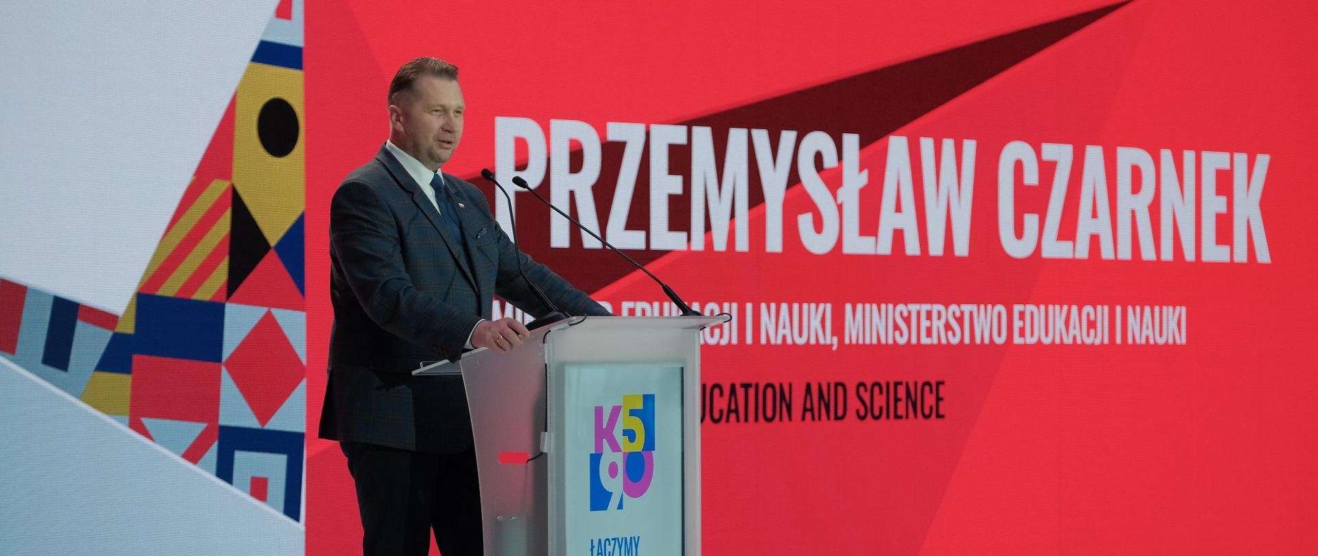 Minister Czarnek stoi na mównicy i mówi do dwóch mikrofonów, za nim czerwona ściana z napisem Przemysław Czarnek.