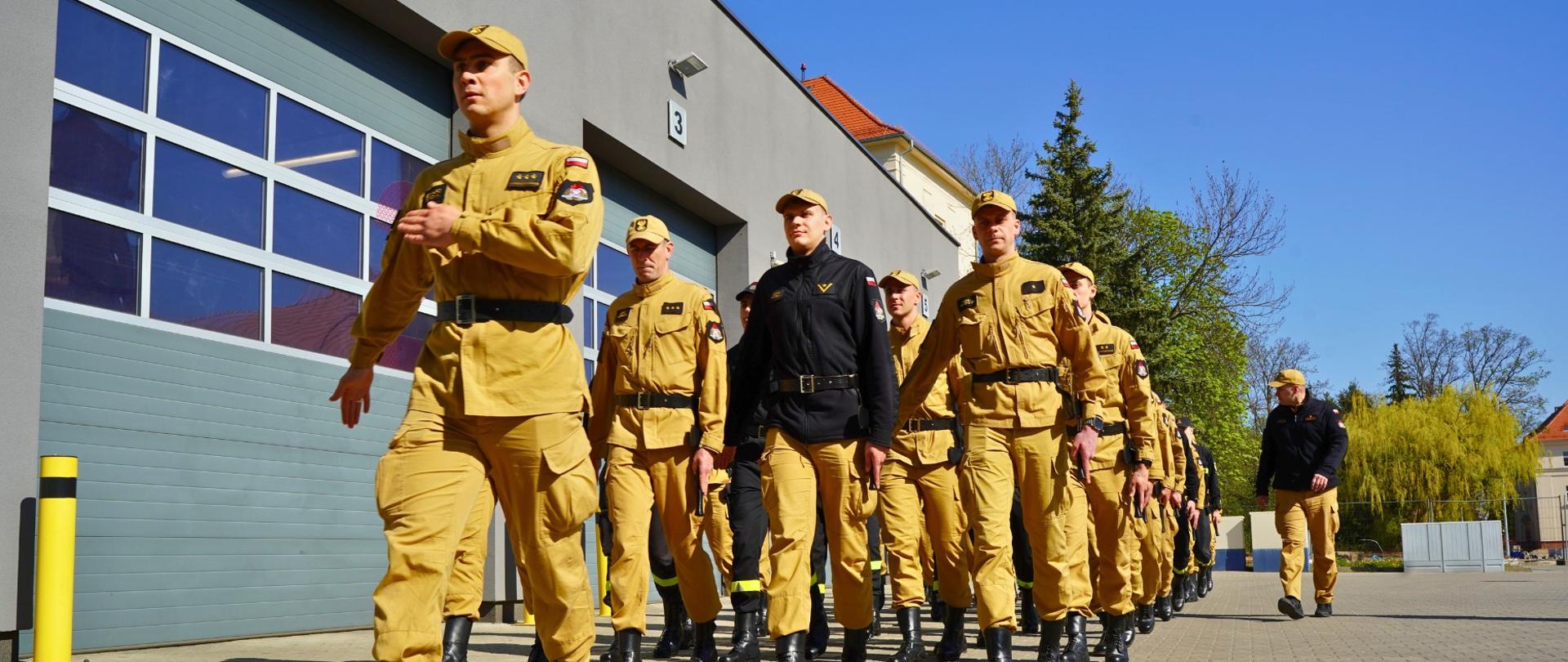 Strażacy ustawieni w pododdział marszowy, w równym umundurowaniu koloru piaskowego, maszerują na placu przed remizą jednostki, ćwicząc do uroczystości z okazji Dnia Strażaka.
