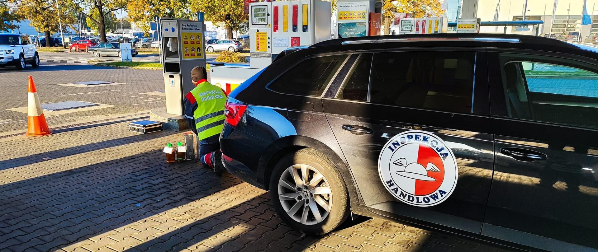 Na pierwszym planie czarny samochód kombi z logiem Inspekcji Handlowej na tylnych drzwiach. Na drugim planie Inspektor Inspekcji Handlowej w trakcie kontroli paliw na stacji benzynowej. W tle infrastruktura stacji paliw. 