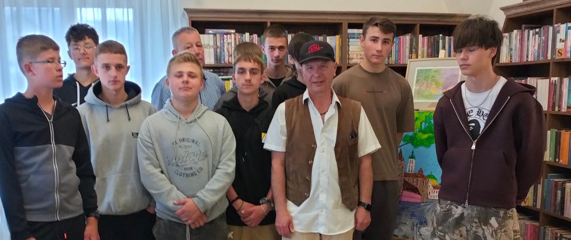 Grupa młodych osób wraz z opiekunem pozuje do wspólnego zdjęcia w jasnym pomieszczeniu biblioteki. W tle znajdują się regały wypełnione książkami oraz obrazy prezentowane na ścianach i sztalugach. Wnętrze wyróżniają wysokie, łukowe sklepienia oraz przestronna aranżacja.