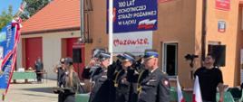Trzech strażaków w mundurach salutuje w stronę sztandaru jednostki straży pożarnej. Tuż za nimi stoją biało-czerwone flagi. W tle budynek remizy strażackiej z napisem "100 lat Ochotniczej Straży Pożarnej w Łącku. 