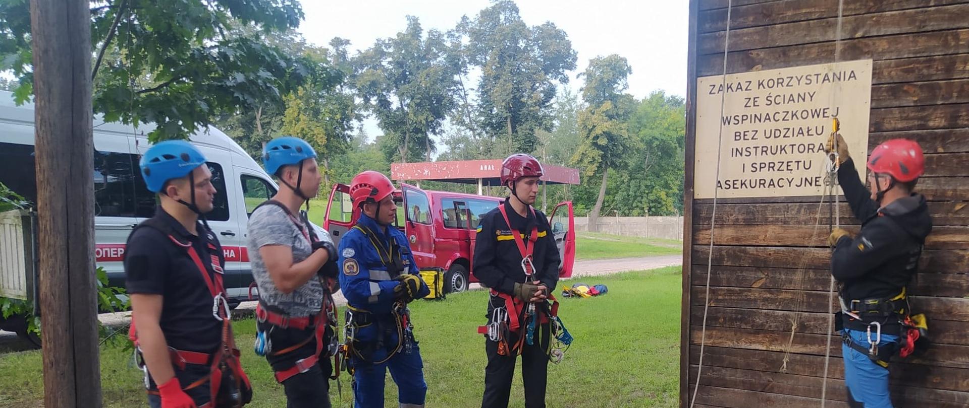 Podczas szkolenia 4 uczestników stoi po lewej stronie a instruktor po prawej przy ścianie drewnianej ścianki wspinaczkowej, na której widnieje tablica z napisem ZAKAZ KORZYSTANIA ZE ŚCIANY WSPINACZKOWEJ BEZ UDZIAŁU INSTRUKTORA I SPRZĘTU ASEKURACYJNEGO. W tle samochody ratownicze i drzewa