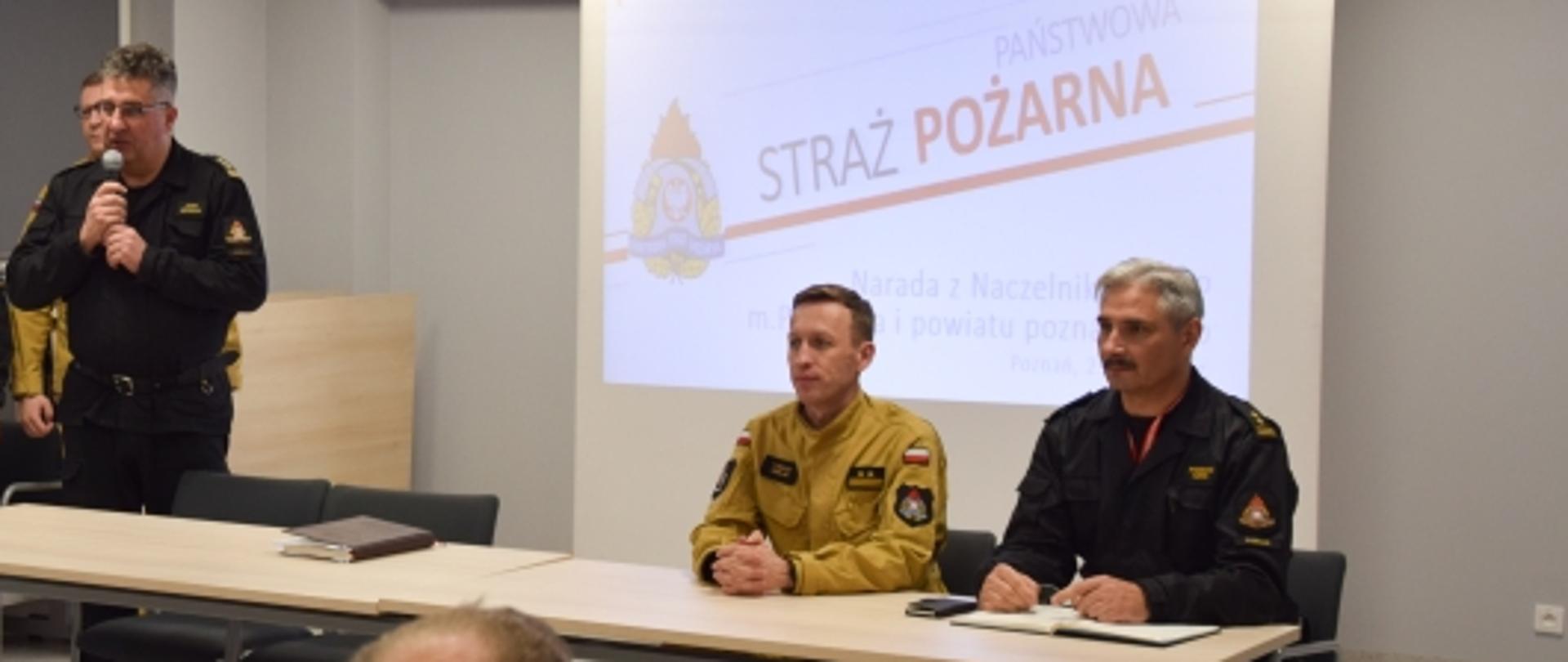 dwóch strażaków siedzi przy stole, dwóch obok jy ekran z logo psp oraz napisem straż pożarna