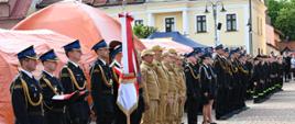 Wzdłuż placu stoi długi szereg funkcjonariuszy w różnych mundurach: galowych czarnych z czapkami rogatywkami oraz służbowych w kolorze piaskowym. Na pierwszym planie widoczny jest poczet sztandarowy oraz strażacy trzymający złożoną flagę państwową. W tle znajdują się duże pomarańczowe namioty oraz fragmenty zabytkowej zabudowy miejskiej.