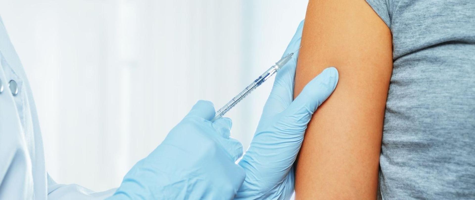 po prawej stronie na jasnym tle, na środku ręce w rękawiczkach ze strzykawką, osoba jest szczepiona