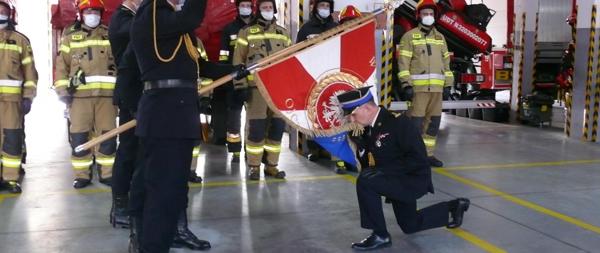 Zdający obowiązki komendanta st. bryg. Adam Dominikowski, klęcząc całuje sztandar jednostki. Sztandar pochyla poczet sztandarowy. Komendant i poczet są w umundurowaniu galowym, zaś w tle towarzyszy im załoga, ubrana w ubrania specjalne. Miejscem uroczystości jest garaż miejscowej jednostki.