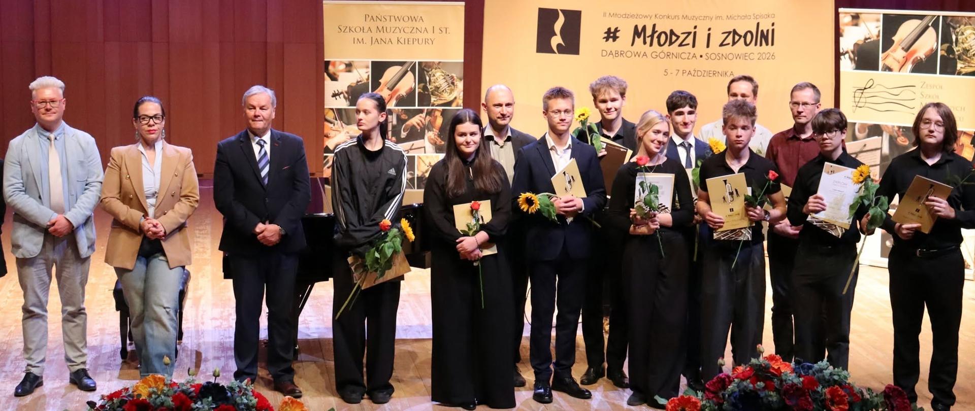 Uczniowie i jury stoją na scenie z kwiatami i dyplomami, w tle duże banery i kotara