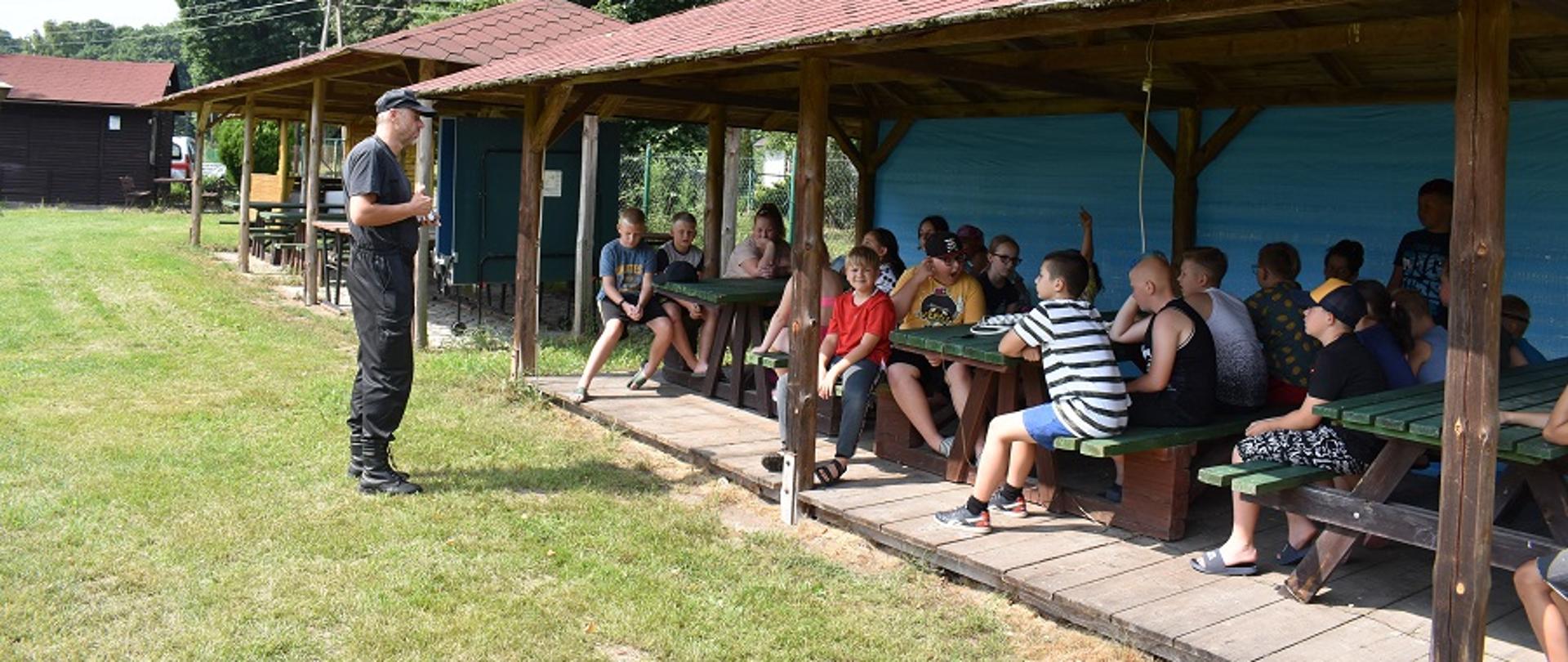 Funkcjonariusz PSP ubrany w mundur dowódczo-sztabowy bez bluzy prowadzi pogadankę dla grupy dzieci siedzący na drewnianych ławkach znajdujących się pod wiatą.