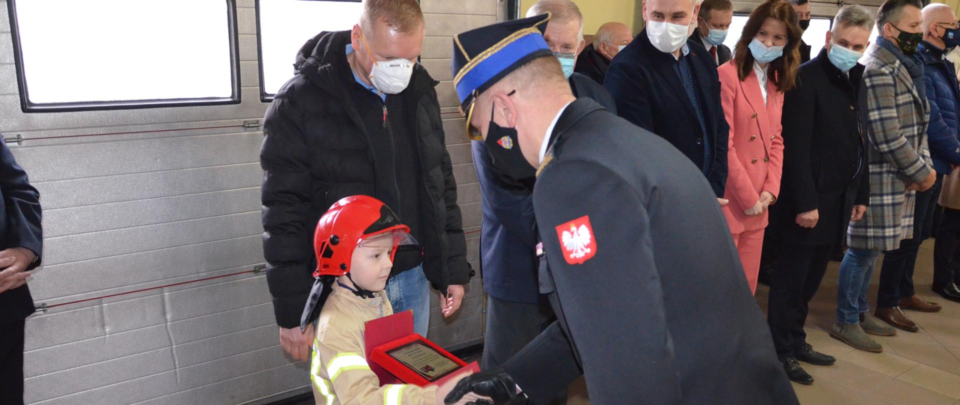 Zdjęcie przedstawia starszego brygadiera Zbigniewa Łyszkowicza Zastępcę Komendanta Wojewódzkiego Państwowej Straży Pożarnej w Łodzi gratulującemu chłopcowi w stroju strażaka, w tle inne osoby 