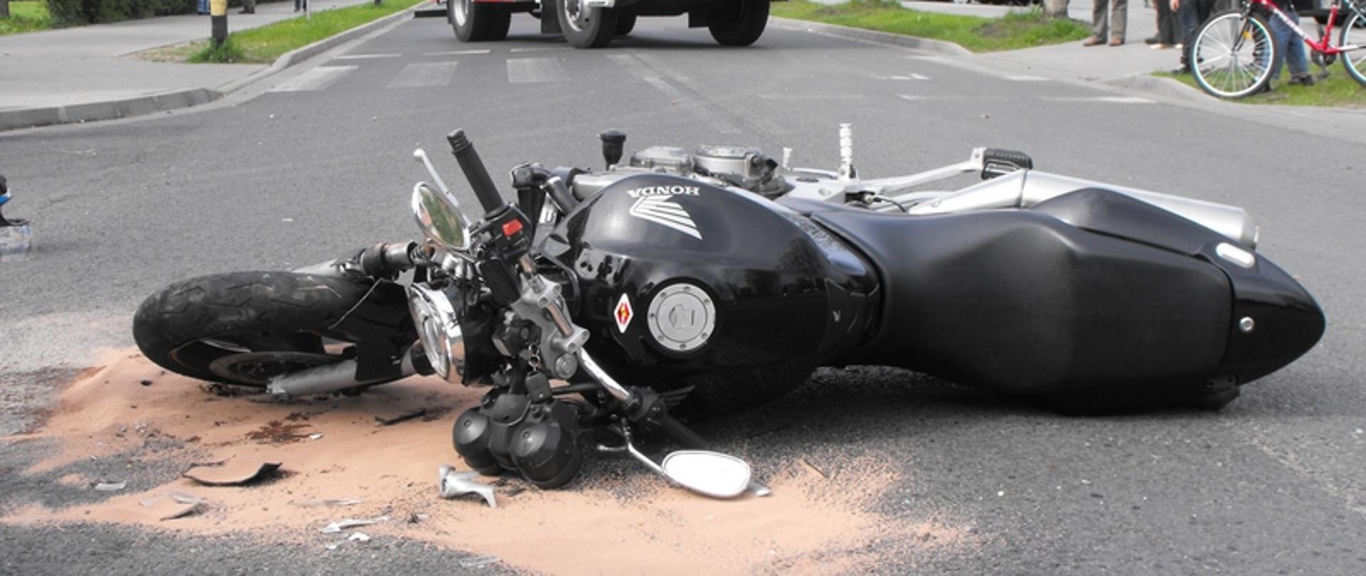 Zdjęcie przedstawia rozbity motocykl na drodze w tle samochód gaśniczy