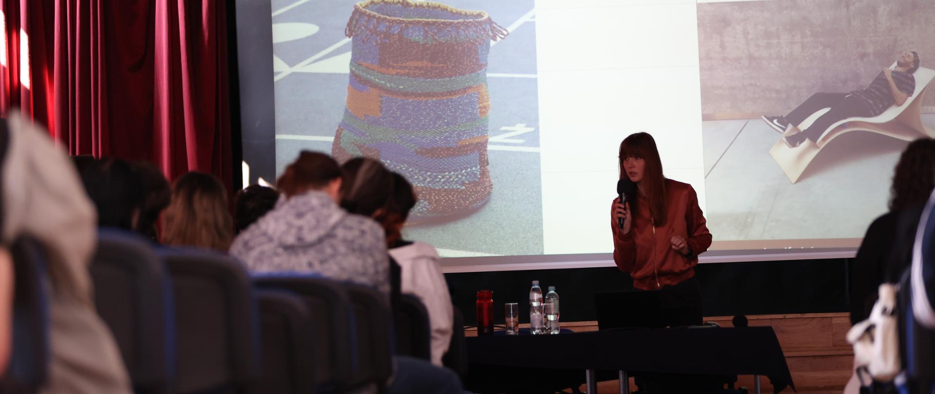 Na zdjęciu młoda kobieta Patrycja Mikołajczak przemawia do uczniów w auli PLSP w Lublinie. Stoi i trzyma w rękach mikrofon na tle ekranu, na którym wyświetlany jest slajd prezentujący jej uczelnię.