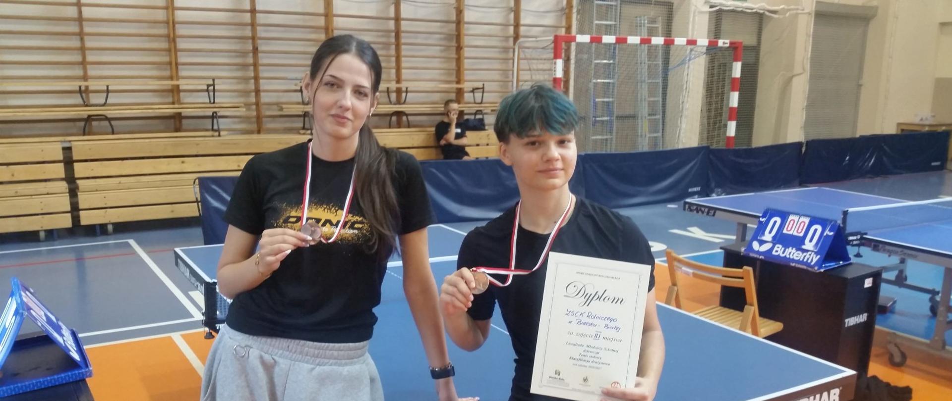 Dwie osoby stoją przy stole do tenisa stołowego w hali sportowej. Każda ma na szyi medal zawieszony na biało-czerwonej wstążce i trzyma go w dłoni. Osoba po prawej dodatkowo trzyma dyplom. W tle widoczne są kolejne stoły do tenisa stołowego, tablice wyników, bramka sportowa oraz drewniane trybuny.