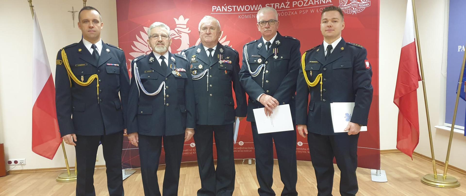Na tle czerwonego baneru stoją w szeregu awansowani i odznaczeni strażacy.