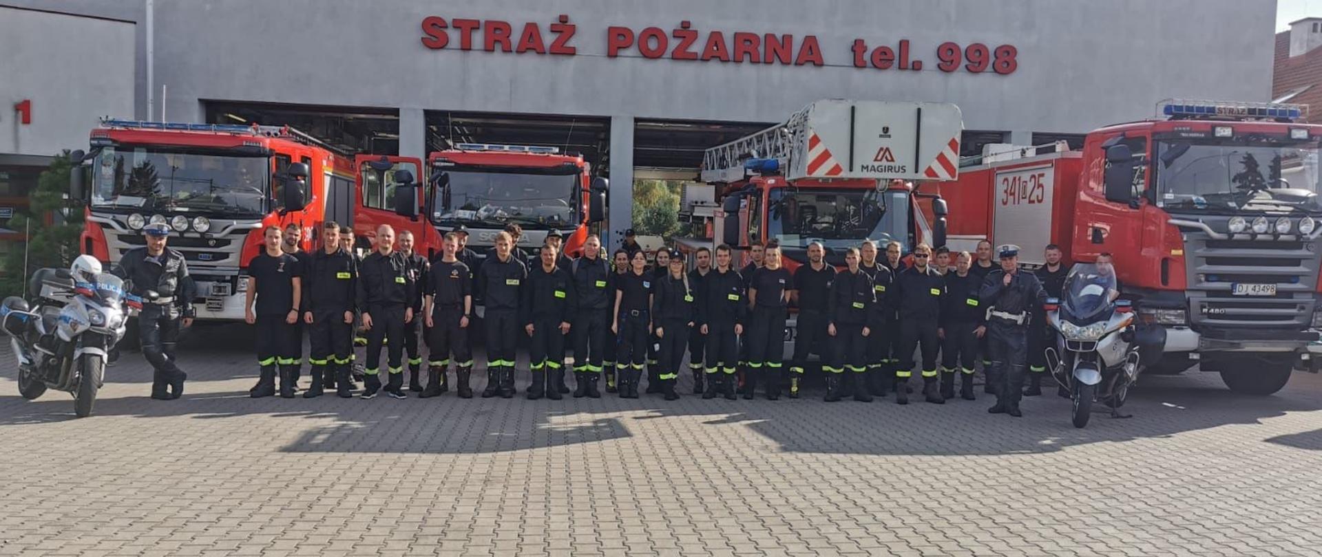 Na zdjęciu widać strażaków wraz z policjantami. W tle za plecami strażaków stoją samochody strażackie koloru czerwonego. Obok strażaków stoją policjanci wraz z motocyklami na obrzeżach. Nad samochodami strażackimi widnieje napis straż pożarna tel 998. Zdjęcie zrobione jest w dzień na placu jednostki. 