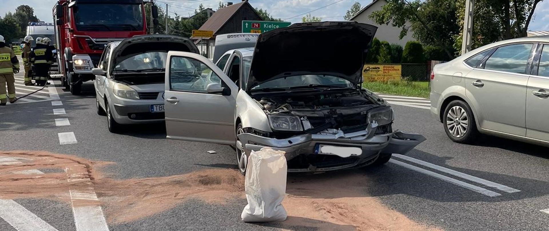 Zdjęcie przedstawia drogę krajową gdzie doszło do wypadku samochodowego. Na pierwszym planie jedno z aut biorących udział w zdarzeniu. Przed nim rozsypany Damolin usuwający plamy płynów. Za nim drugi ze zniszczonych aut, a w tle samochód pożarniczy, przy którym stoją strażacy. Sąsiednim pasem przejeżdżają przypadkowe samochody.