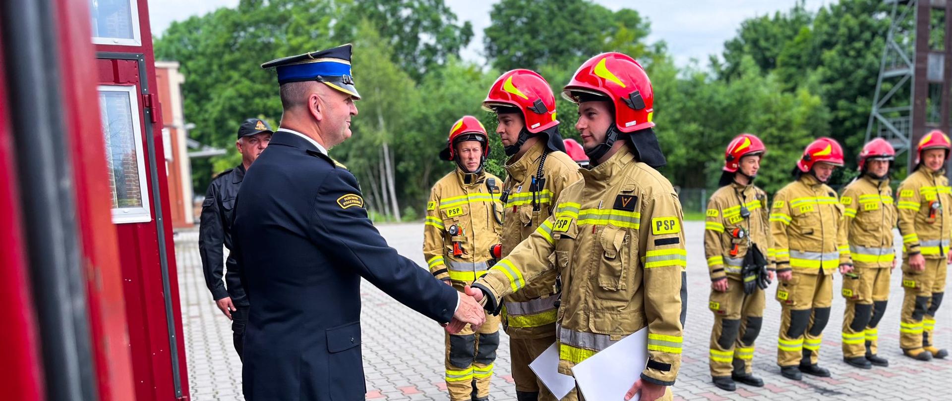 Komendant powiatowy Państwowej Straży Pożarnej w Ostrzeszowie ściska dłoń strażakowi po wręczeniu wyróżnienia. Na drugim planie strażacy w ubraniach specjalnych. W tle drzewa. Zdjęcie wykonane na placu wewnętrznym jednostki.