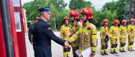 Komendant powiatowy Państwowej Straży Pożarnej w Ostrzeszowie ściska dłoń strażakowi po wręczeniu wyróżnienia. Na drugim planie strażacy w ubraniach specjalnych. W tle drzewa. Zdjęcie wykonane na placu wewnętrznym jednostki.