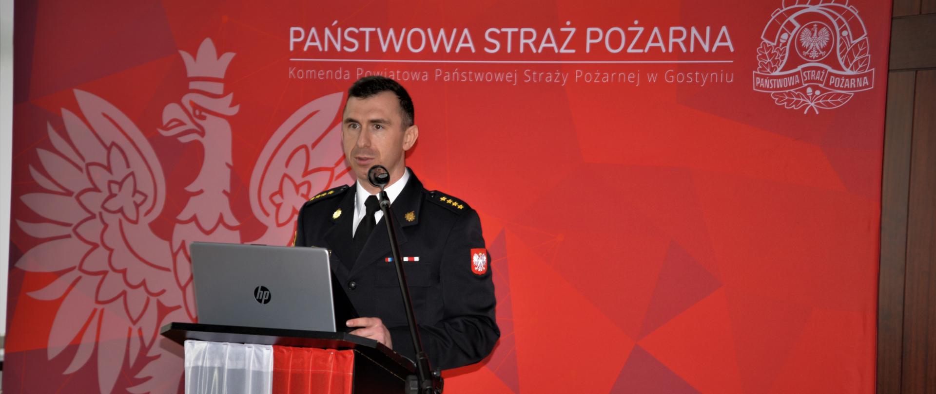 Zdjęcie przedstawia Komendanta Powiatowego PSP w Gostyniu podczas przedstawiania prezentacji multimedialnego. Na mównicy położony jest laptop.